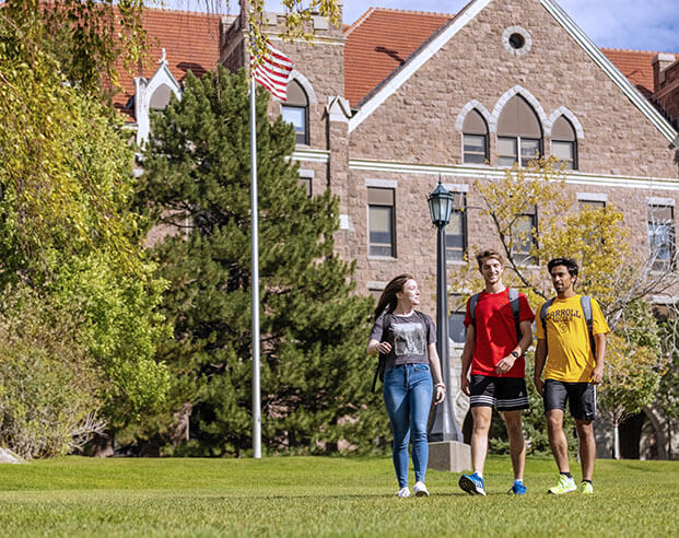 Three students strolling along the grass on a sunny day in front of a beautiful brick building