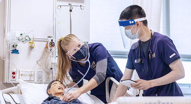 Two students wearing PPE while checking the heart rate of a dummy in a hospital bed