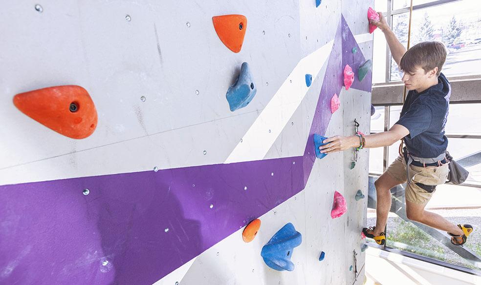 Student stretches his arm while trying to grab a rock hold in the climbing gym